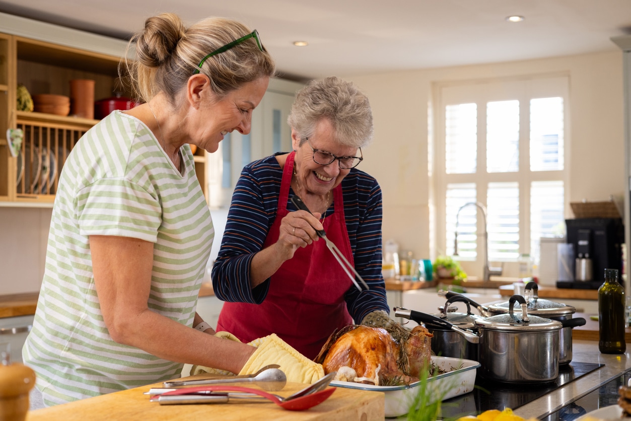 Waist-up shot of a senior lady with her daughter basting a fresh turkey fresh out of an oven.